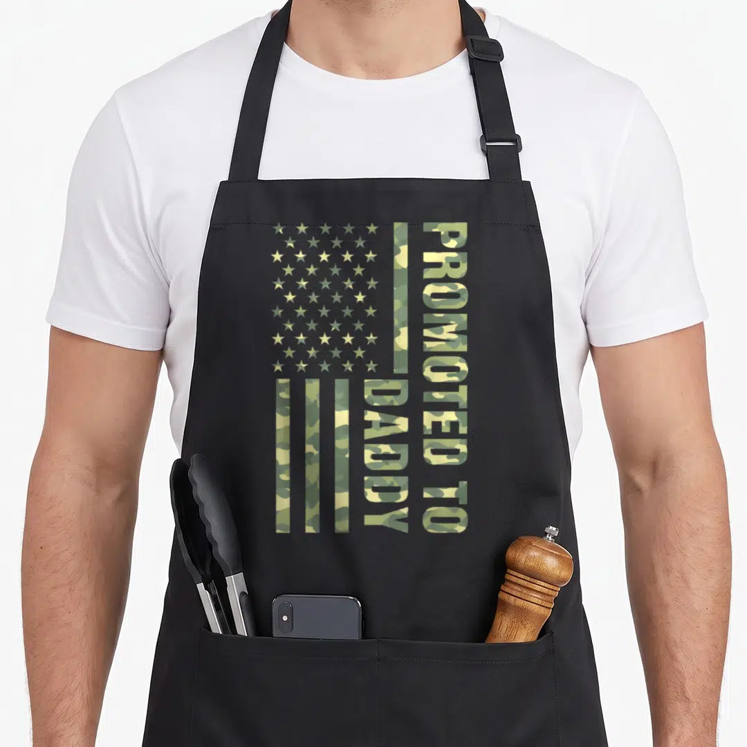 Promoted father day apron - Camouflage USA flag with vertical PROMOTED TO DADDY text in olive and tan — shown on a black cotton canvas apron against a white background