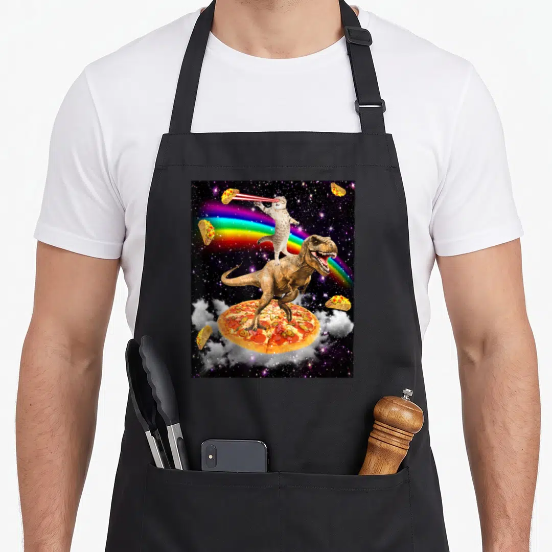 Laser taco apron - T-Rex on pizza with laser cat rainbow and floating tacos in space — shown on a black cotton canvas apron against a white background