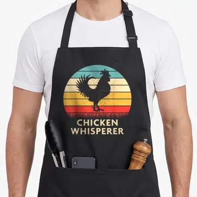 Cute whisperer chicken apron - Retro rooster silhouette over striped sun with CHICKEN WHISPERER text in teal yellow red — shown on a black cotton canvas apron against a white background