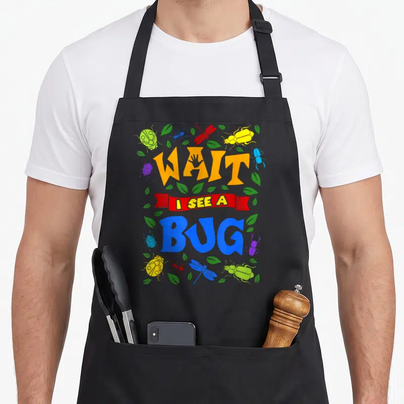 Wait pull girl apron - Colorful cartoon bugs surround stacked text reading WAIT I SEE A BUG with a handprint — shown on a black cotton canvas apron against a white background