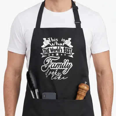 Funny family gathering apron - White skeleton hands holding a banner reading This is What THE World's BEST Family looks — shown on a black cotton canvas apron against a white background