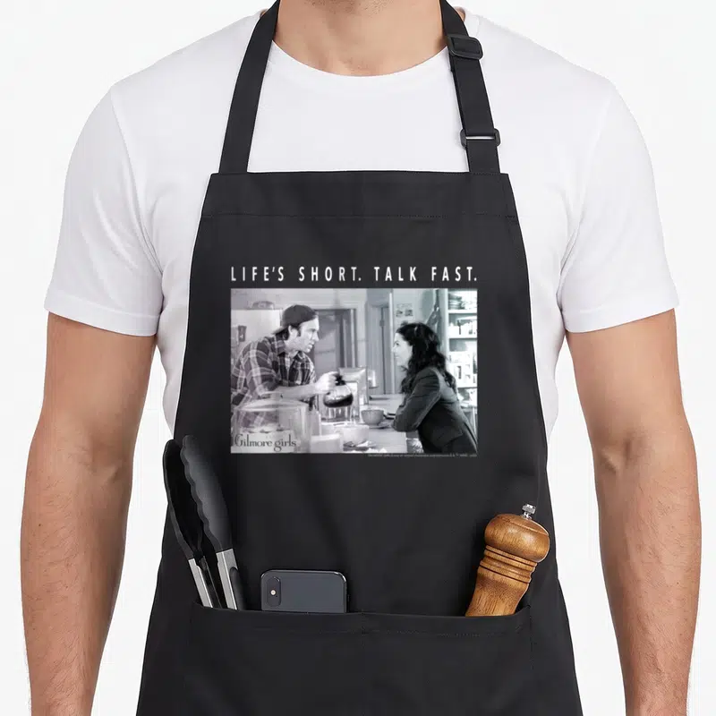 Luke gilmore apron - LIFE'S SHORT. TALK FAST. text above grayscale photo of people at a counter — shown on a black cotton canvas apron against a white background