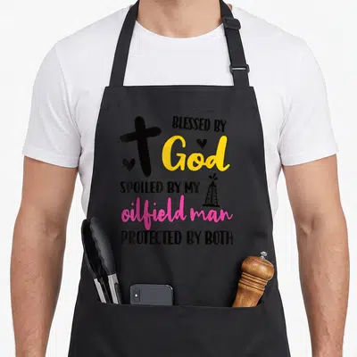 Spoiled oilfield husband wife aprons - Black cross yellow God pink oilfield man text oil derrick two hearts — shown on a black cotton canvas apron against a white background