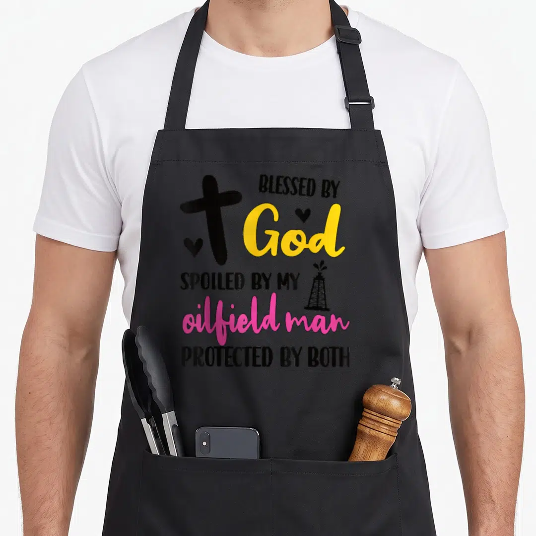 Spoiled oilfield husband wife aprons - Black cross yellow God pink oilfield man text oil derrick two hearts — shown on a black cotton canvas apron against a white background