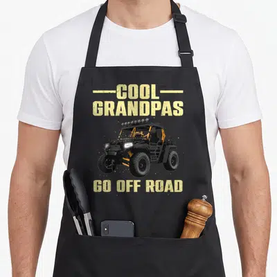 Offroad grandpa apron - Distressed yellow text COOL GRANDPAS GO OFF ROAD above a black and orange UTV illustration — shown on a black cotton canvas apron against a white background