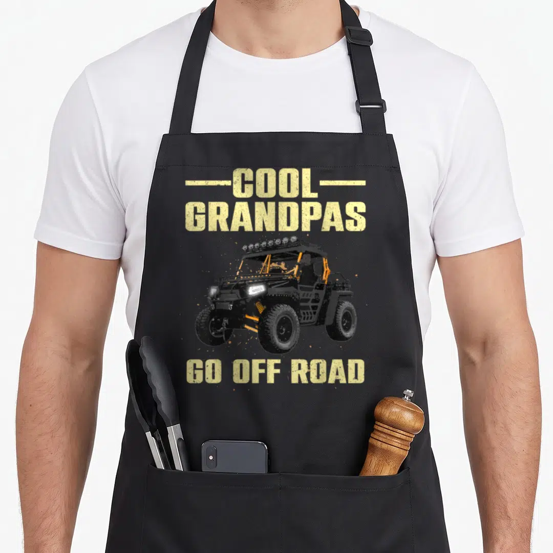 Offroad grandpa apron - Distressed yellow text COOL GRANDPAS GO OFF ROAD above a black and orange UTV illustration — shown on a black cotton canvas apron against a white background