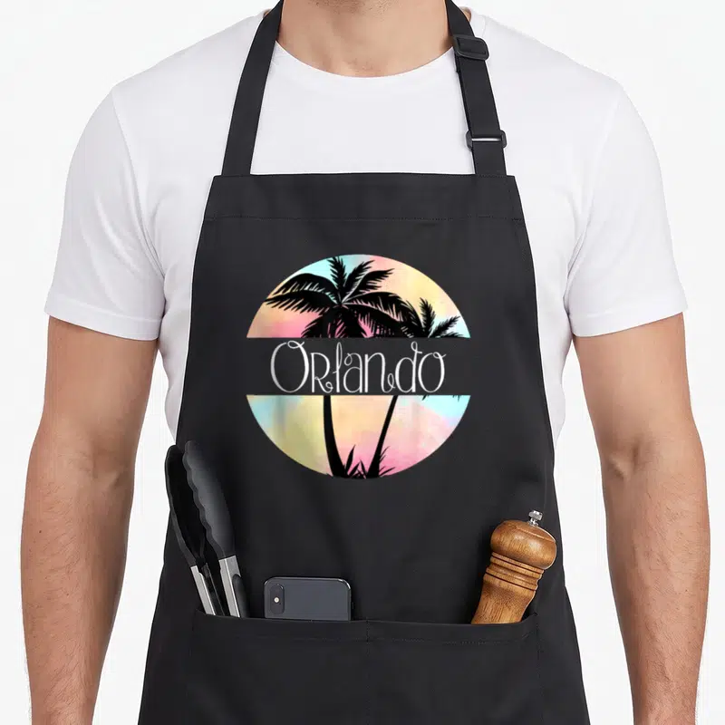 Orlando tree apron - Black palm tree silhouettes on pink yellow blue watercolor circle with white Orlando text — shown on a black cotton canvas apron against a white background