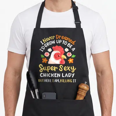 Cute farmer chicken with apron - Cartoon chicken head with flowers and text I Never Dreamed I'd Grow Up To Be A Super — shown on a black cotton canvas apron against a white background