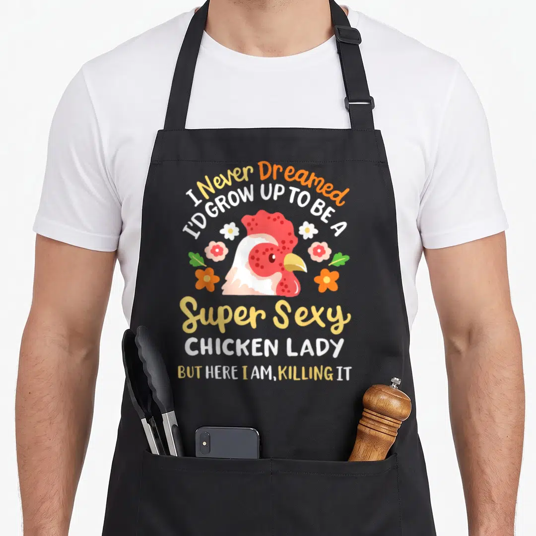 Cute farmer chicken with apron - Cartoon chicken head with flowers and text I Never Dreamed I'd Grow Up To Be A Super — shown on a black cotton canvas apron against a white background