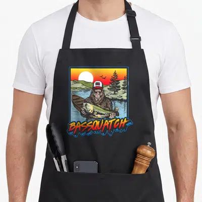 Bassquatch fishing aprons - Cartoon bigfoot holding a fish in a lake scene with BASSQUATCH text below — shown on a black cotton canvas apron against a white background