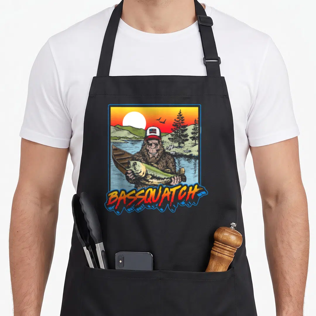 Bassquatch fishing aprons - Cartoon bigfoot holding a fish in a lake scene with BASSQUATCH text below — shown on a black cotton canvas apron against a white background