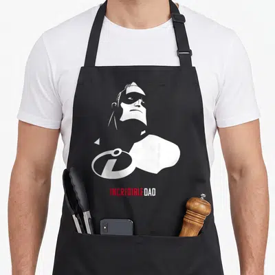 Incredible dad mode apron - White male silhouette above red INCREDIBLE DAD text with overlapping circular logo on grey — shown on a black cotton canvas apron against a white background