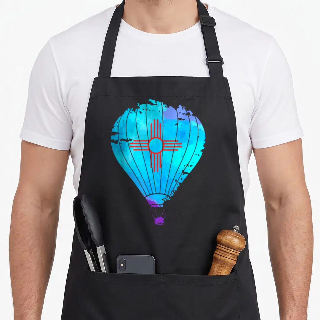 Mexico balloon apron - Watercolor hot air balloon with red Zia sun symbol in cyan, blue, and purple — shown on a black cotton canvas apron against a white background