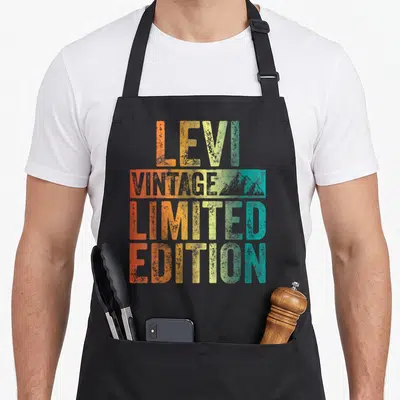 Limited first edition apron - LEVI VINTAGE LIMITED EDITION text in orange yellow green teal with grey mountain graphic — shown on a black cotton canvas apron against a white background