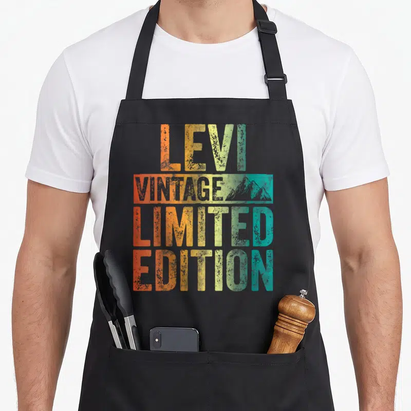 Limited first edition apron - LEVI VINTAGE LIMITED EDITION text in orange yellow green teal with grey mountain graphic — shown on a black cotton canvas apron against a white background