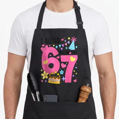 Birthday 7th bow apron - Pink 67 numbers with I WAS NOW I'M text, party hat, cake, and scattered hearts stars flowers — shown on a black cotton canvas apron against a white background
