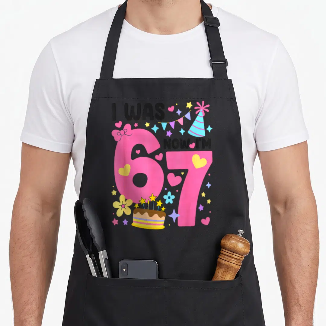 Birthday 7th bow apron - Pink 67 numbers with I WAS NOW I'M text, party hat, cake, and scattered hearts stars flowers — shown on a black cotton canvas apron against a white background