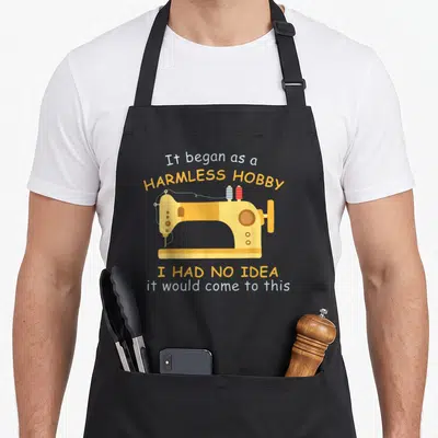 Sewing hobby apron - Yellow cartoon sewing machine with blue and red thread spools and sarcastic quote text — shown on a black cotton canvas apron against a white background