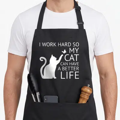 Cute hard cat apron - White cat silhouette reaching for a butterfly next to stacked text I WORK HARD SO MY CAT CAN HAVE — shown on a black cotton canvas apron against a white background