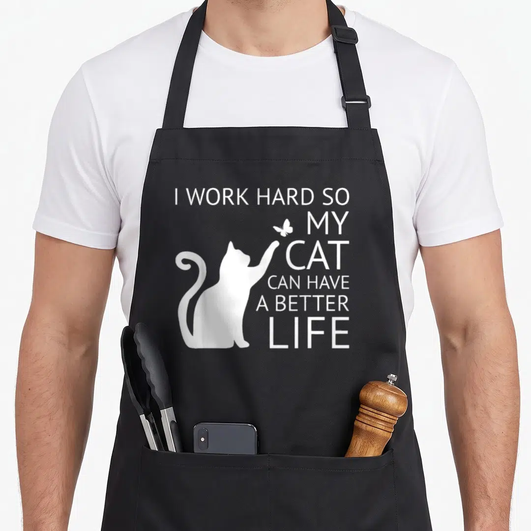 Cute hard cat apron - White cat silhouette reaching for a butterfly next to stacked text I WORK HARD SO MY CAT CAN HAVE — shown on a black cotton canvas apron against a white background