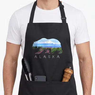 Wildflower bear apron - Bear silhouette filled with snowy mountain, blue water, green plants, and purple flowers above — shown on a black cotton canvas apron against a white background