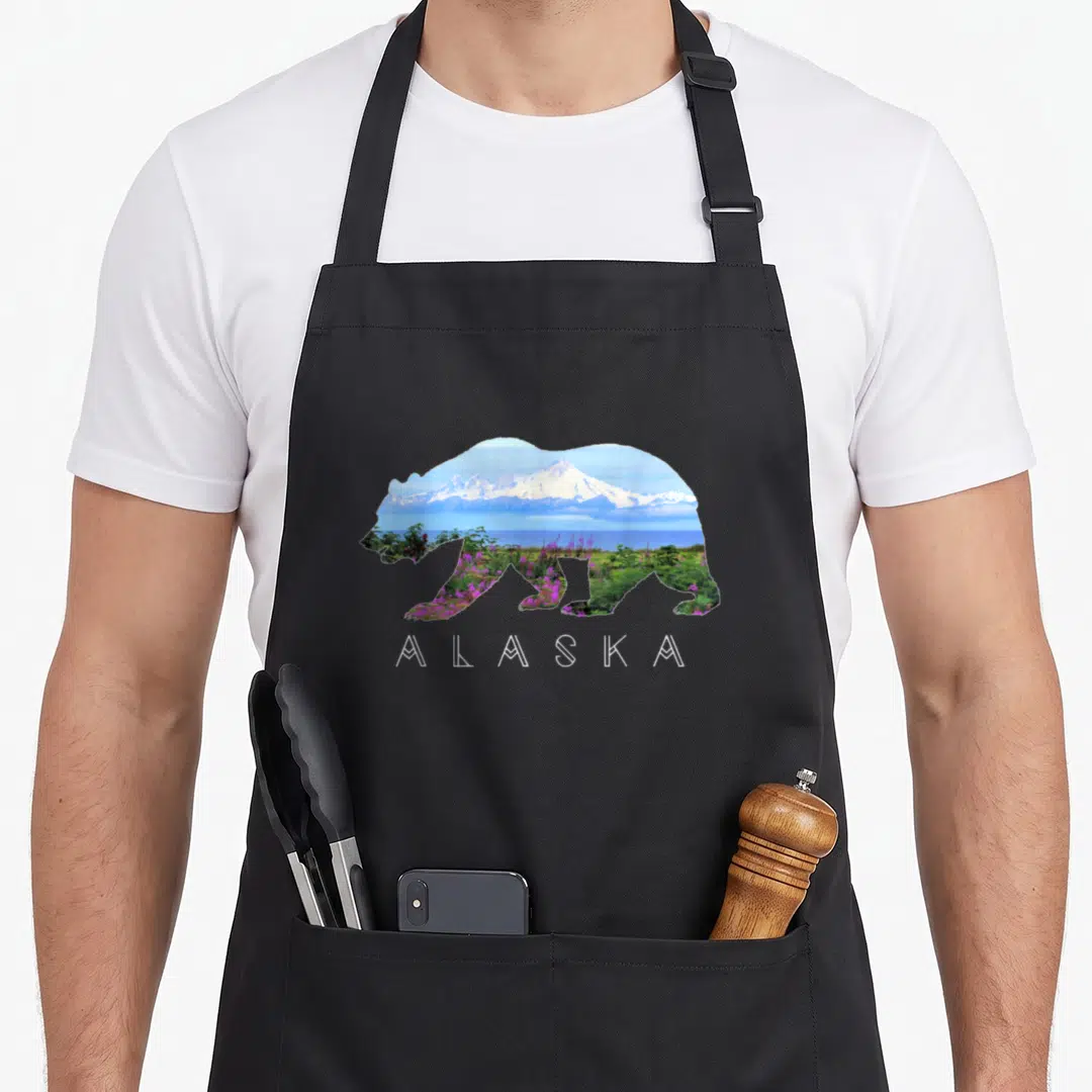 Wildflower bear apron - Bear silhouette filled with snowy mountain, blue water, green plants, and purple flowers above — shown on a black cotton canvas apron against a white background