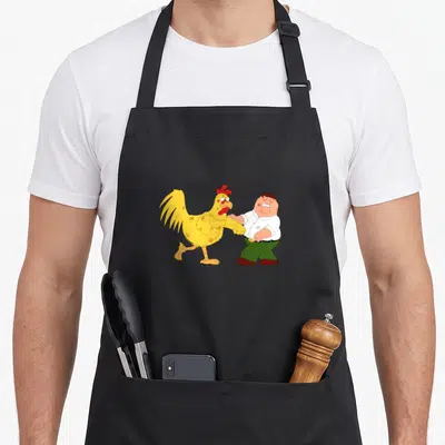 Fighting chicken apron - Cartoon yellow chicken and man in white shirt facing each other in a fight pose — shown on a black cotton canvas apron against a white background