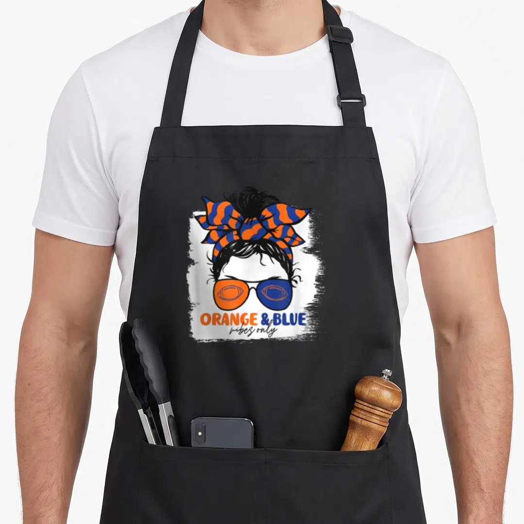 Blue messy aprons - Cartoon woman with striped bow and football sunglasses above orange and blue text — shown on a black cotton canvas apron against a white background
