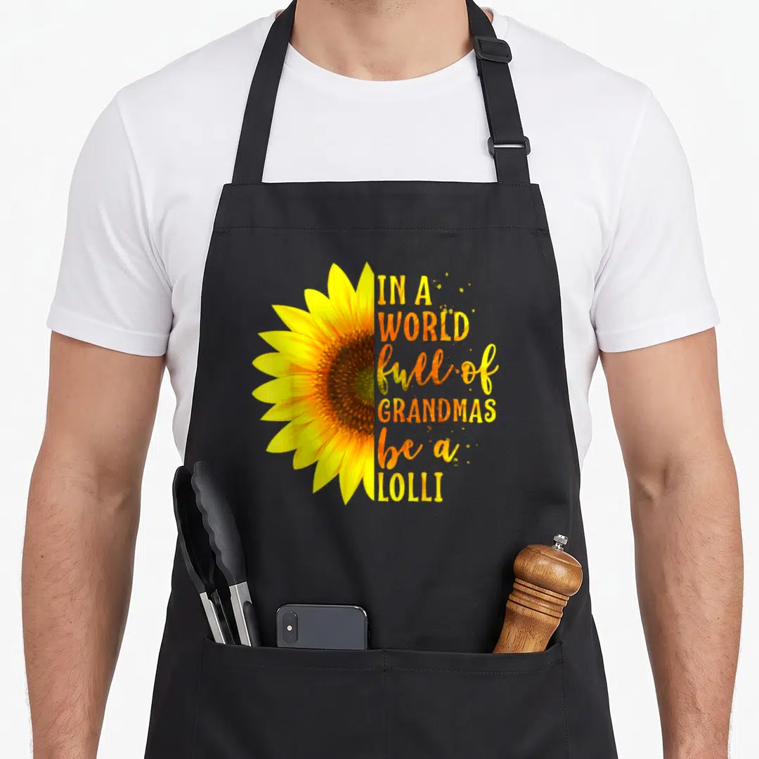 Grandmas mother's day aprons - Half sunflower in yellow and brown beside stacked text reading IN A WORLD full — shown on a black cotton canvas apron against a white background