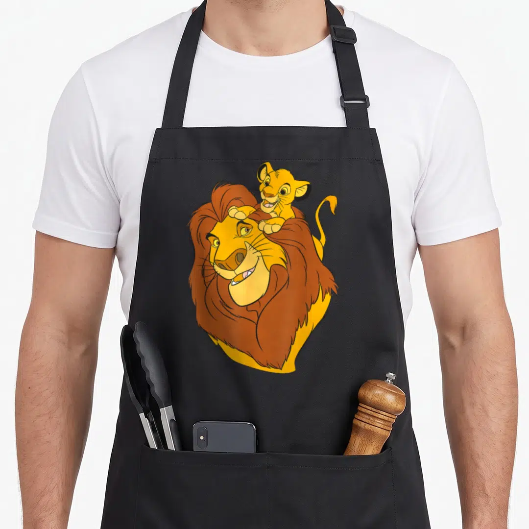 Simba dad and son aprons - Cartoon adult lion with small cub perched on head in yellow and brown — shown on a black cotton canvas apron against a white background