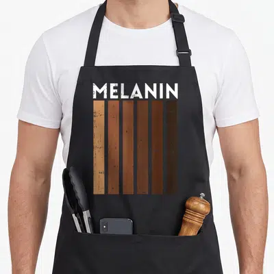 Shade african print apron - MELANIN text above six vertical bars ranging from light tan to dark brown with distressed — shown on a black cotton canvas apron against a white background