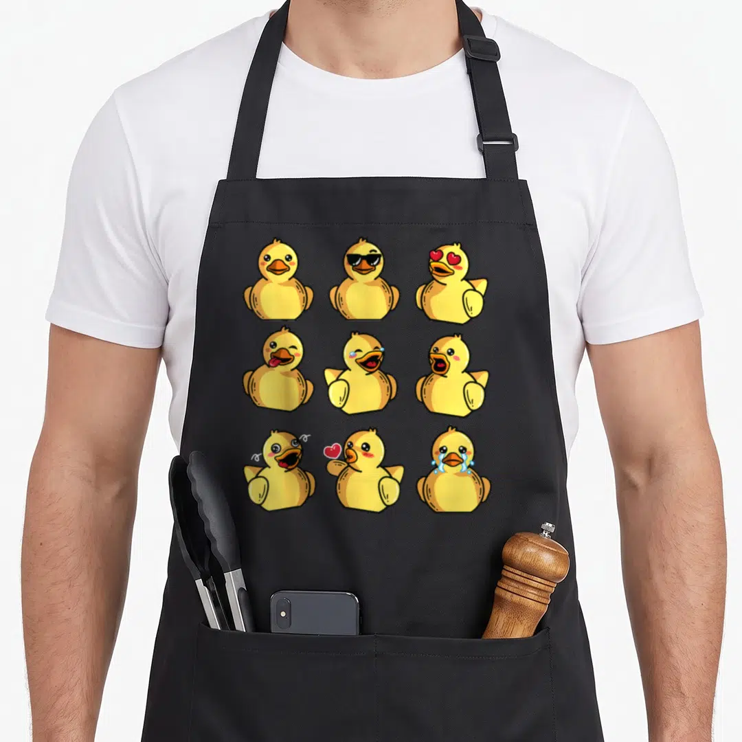 Bath duck apron - Nine yellow cartoon ducks with different faces including sunglasses, hearts, and tears in a grid — shown on a black cotton canvas apron against a white background