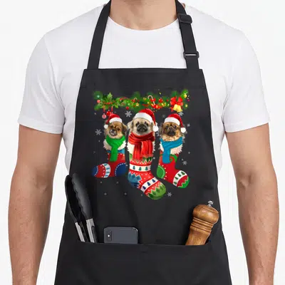 Three tibetan apron - Three dogs in Christmas stockings with Santa hats and scarves under a pine garland with snowflakes — shown on a black cotton canvas apron against a white background