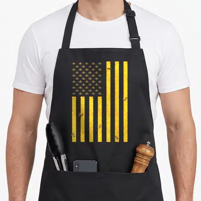 Honeycomb american flag apron - USA flag shape with yellow honeycomb stripes and a grid of cartoon bees filling — shown on a black cotton canvas apron against a white background