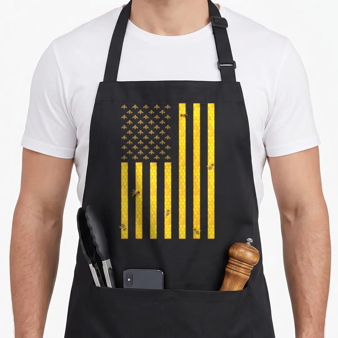 Honeycomb american flag apron - USA flag shape with yellow honeycomb stripes and a grid of cartoon bees filling — shown on a black cotton canvas apron against a white background