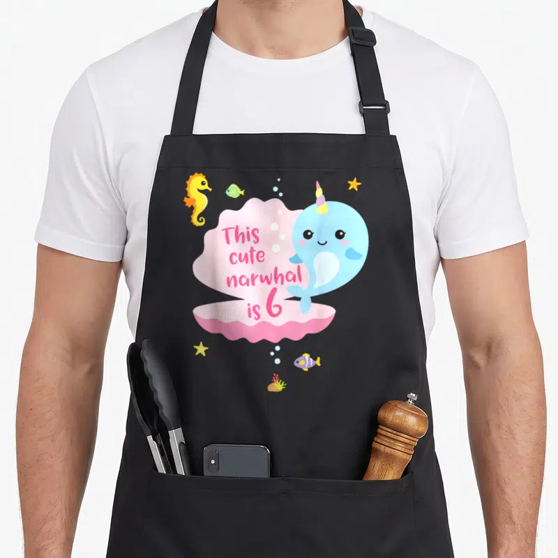 Sea birthday apron - Light blue narwhal next to pink cloud shape with text This cute narwhal is 6 surrounded by sea — shown on a black cotton canvas apron against a white background