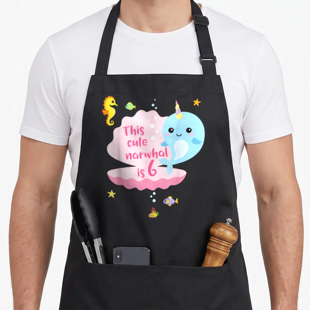 Sea birthday apron - Light blue narwhal next to pink cloud shape with text This cute narwhal is 6 surrounded by sea — shown on a black cotton canvas apron against a white background