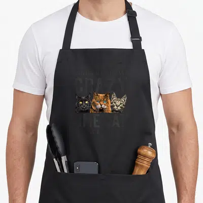 Wife crazy cat lady apron - Three cat heads black orange grey with distressed text DRIVING MY HUSBAND CRAZY ONE CAT — shown on a black cotton canvas apron against a white background