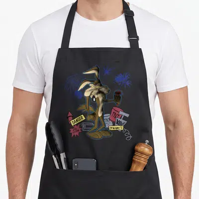 Coyote e.a apron - Wile E. Coyote holding lit fuse with TNT cans and danger signs in brown and blue — shown on a black cotton canvas apron against a white background