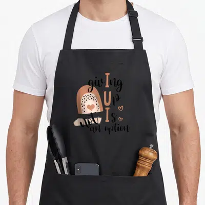 Giving rainbow apron - Stylized tan rainbow with heart and giving up is not an option text — shown on a black cotton canvas apron against a white background