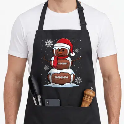 Merry snowman apron - Cartoon snowman made of three brown footballs wearing red hat and scarf with white snowflakes — shown on a black cotton canvas apron against a white background
