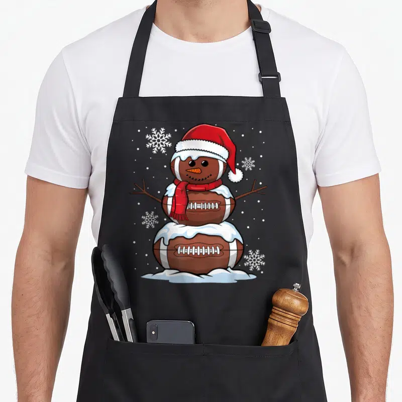 Merry snowman apron - Cartoon snowman made of three brown footballs wearing red hat and scarf with white snowflakes — shown on a black cotton canvas apron against a white background