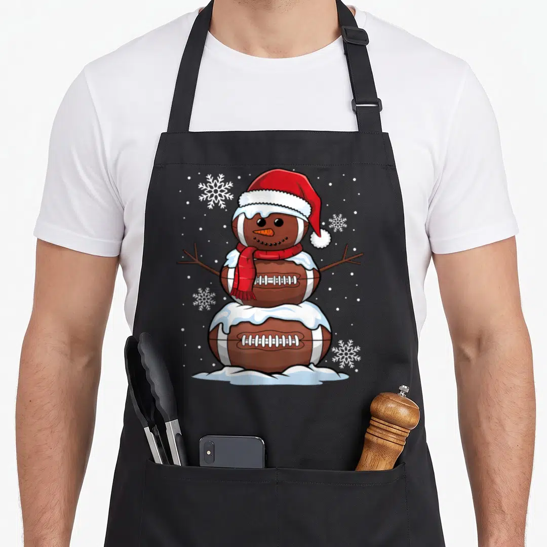 Merry snowman apron - Cartoon snowman made of three brown footballs wearing red hat and scarf with white snowflakes — shown on a black cotton canvas apron against a white background