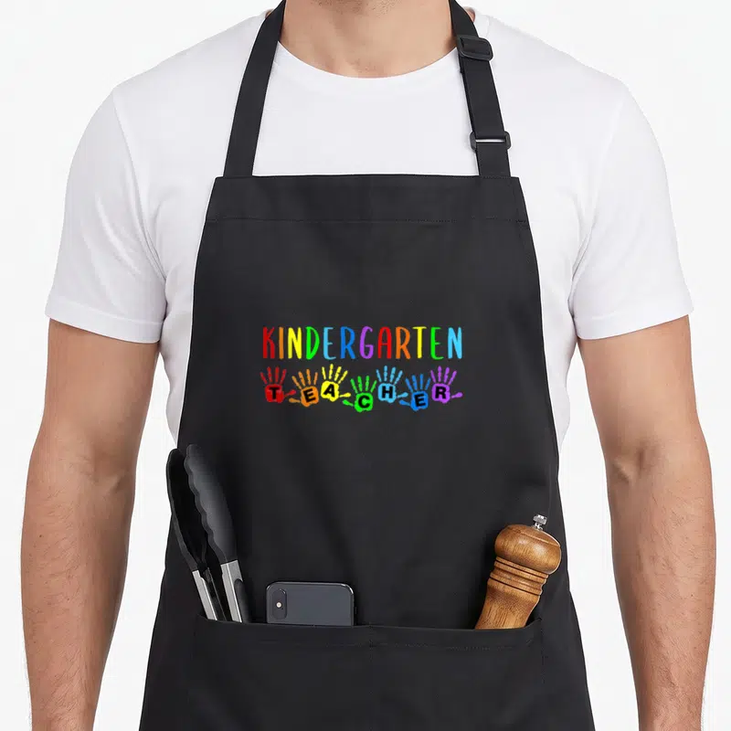 Handprint kindergarten teacher apron - Rainbow text KINDERGARTEN above six colorful handprints spelling TEACHER — shown on a black cotton canvas apron against a white background