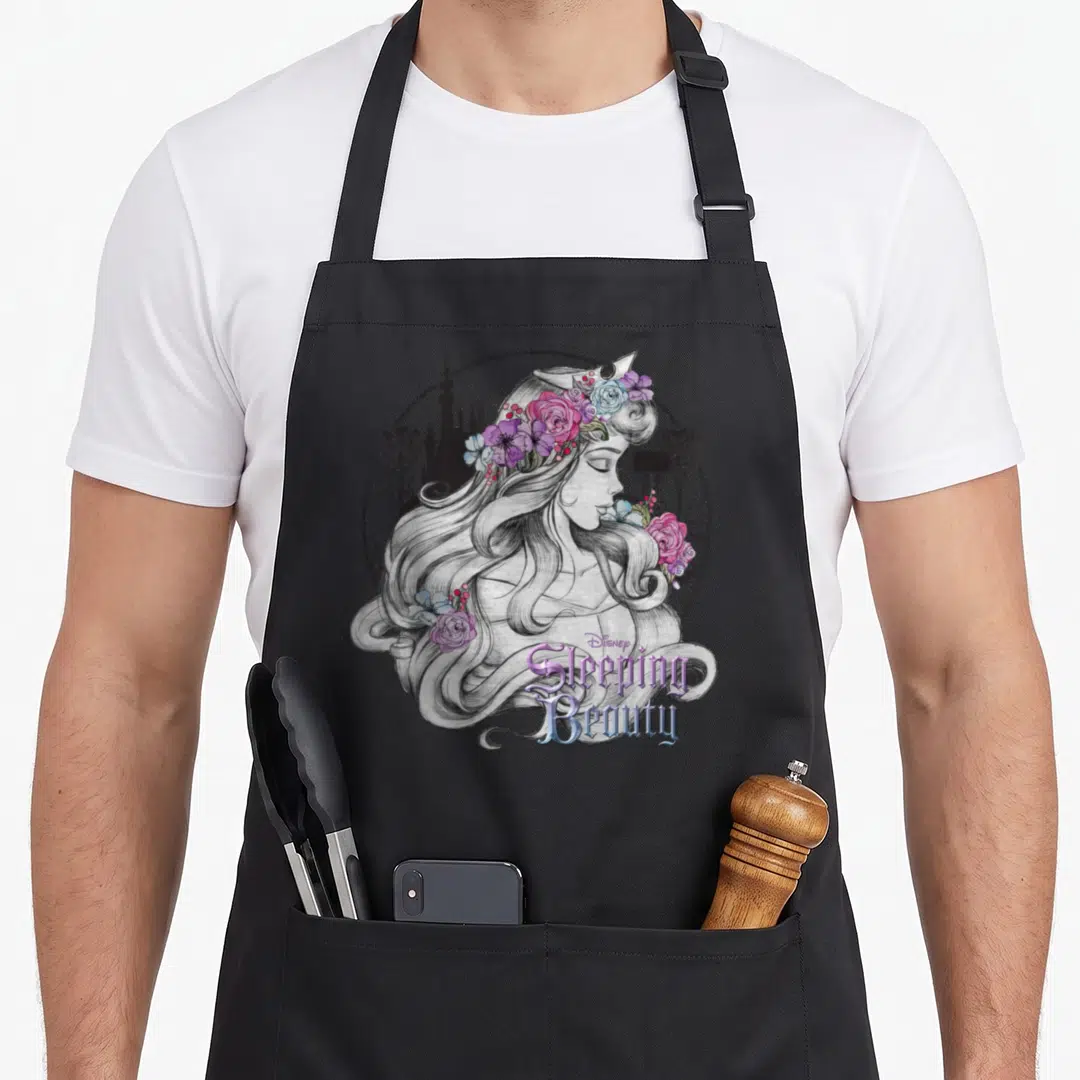 Floral beauty apron - Sketched woman profile with pink purple flower crown and Sleeping Beauty text over castle — shown on a black cotton canvas apron against a white background