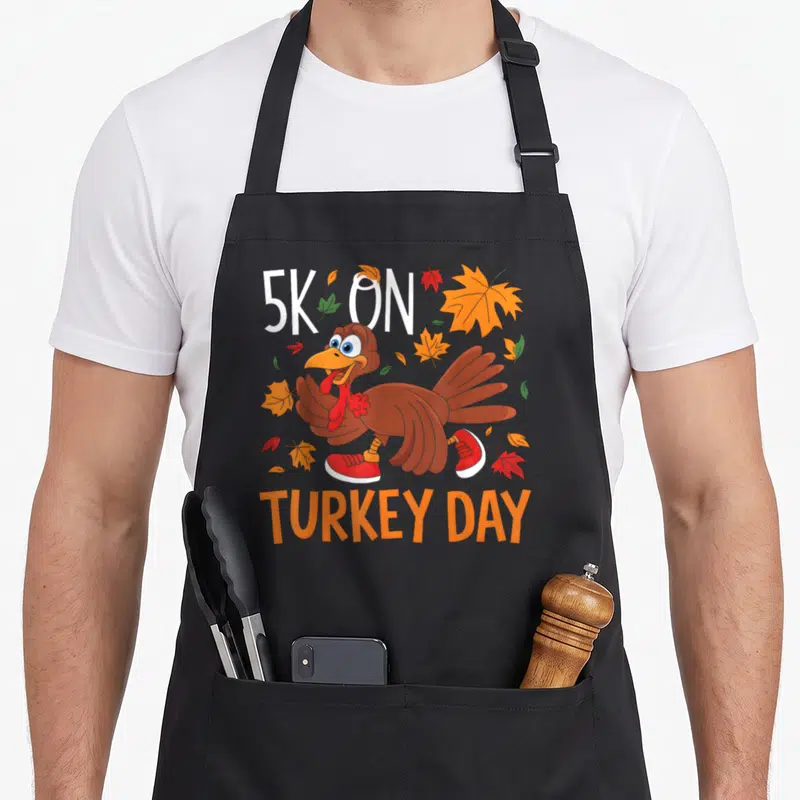 Trot thanksgiving day aprons - Cartoon turkey in red sneakers surrounded by fall leaves with 5K ON TURKEY DAY text — shown on a black cotton canvas apron against a white background