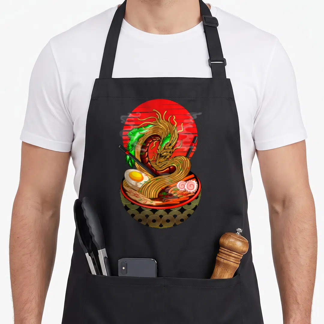 Asian kawaii aprons - Realistic ramen bowl with golden dragon, red sun, black chopsticks, and green garnish — shown on a black cotton canvas apron against a white background