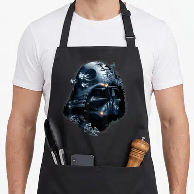 Helmet general apron - Darth Vader helmet formed by space battle collage with Death Star and TIE fighters in black blue — shown on a black cotton canvas apron against a white background