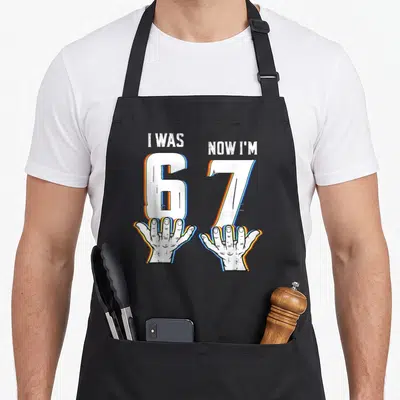 Six meme apron - I WAS 6 NOW I'M 7 text held by two open hands with blue and orange outlines — shown on a black cotton canvas apron against a white background