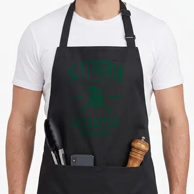 Quidditch potter apron - Dark green coiled snake crossed broomsticks golden snitch SLYTHERIN text distressed print — shown on a black cotton canvas apron against a white background